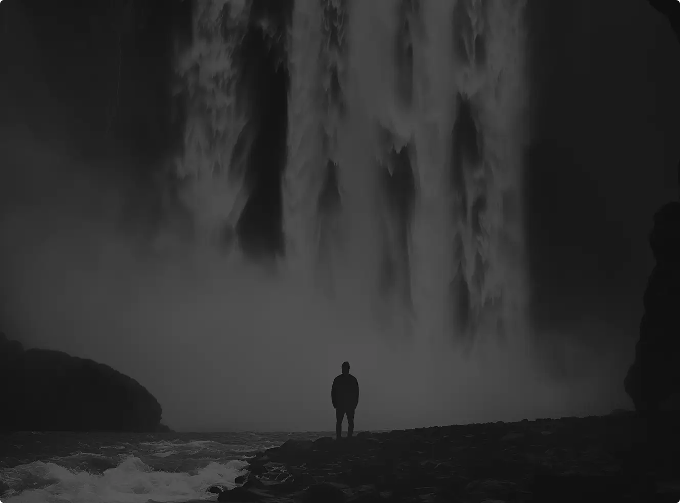 Silhouette of a person standing near a large waterfall with mist and rushing water.