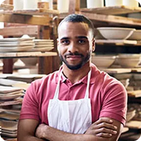 Smiling man in pink shirt and white apron with folded arms standing in pottery workshop.