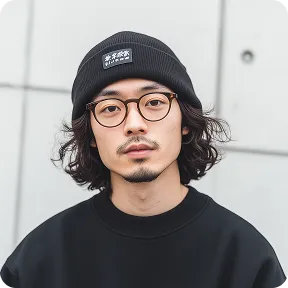 Young man with curly hair, glasses, and a black beanie in a black sweater against a gray wall.