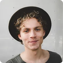 Young man with curly blond hair wearing a black hat smiling softly against a light background.
