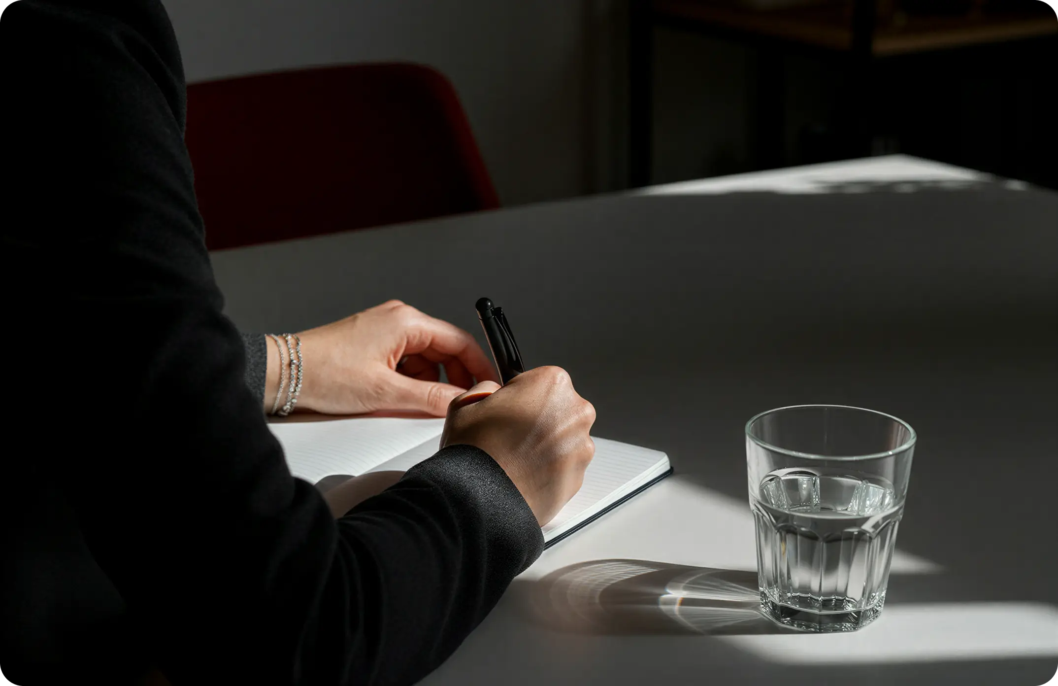 Person writing in a notebook with a pen beside a glass of water on a white table in soft light.